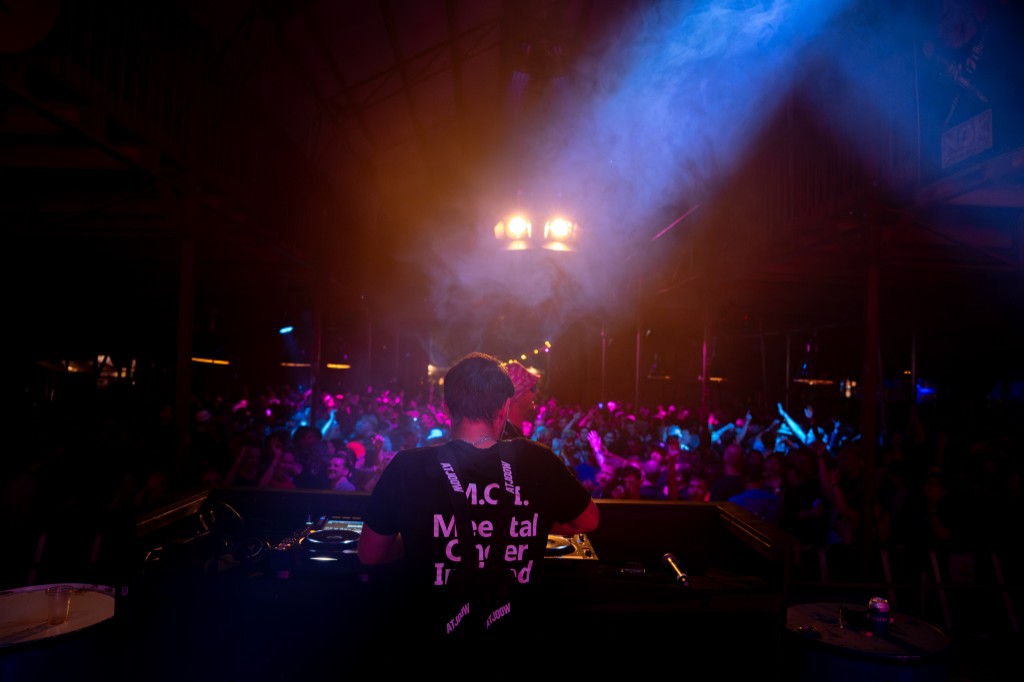 DJ from behind at the Pioneer decks facing a packed crowd under orange stage light