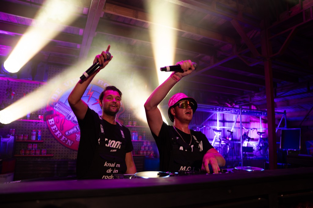 Both Atjoow performers behind the DJ booth with microphones raised under purple light