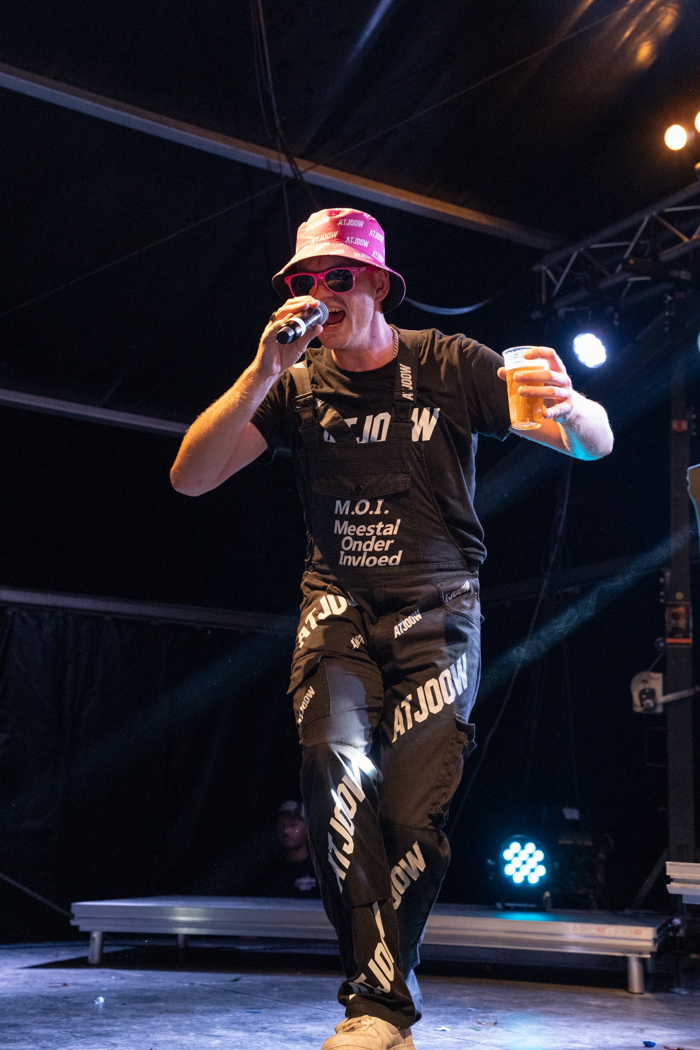 Artist in a pink hat holding a microphone and a drink on stage